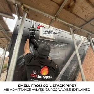 A close-up view of a BDS Services engineer working at height on scaffolding, fitting a specialized black Durgo Air Admittance Valve (AAV) onto a vertical uPVC soil vent pipe stack on a house with grey cladding. The image includes text overlays identifying the components and a title for a blog post about drain smells.